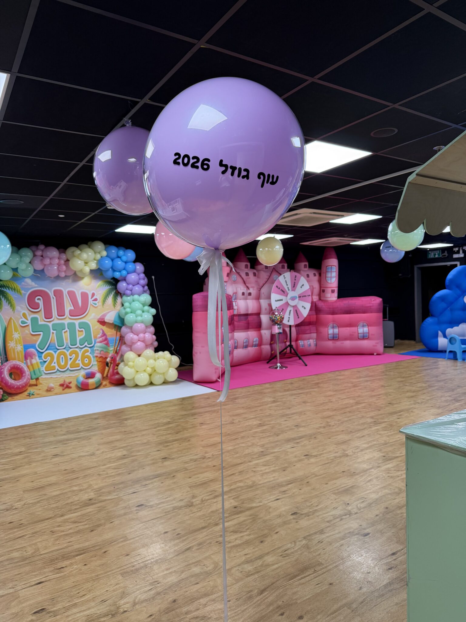 מבט רחב על מתחם עוף גוזל — קיר צילום ממותג, טירה ורודה מתנפחת, גלגל מזל ועמוד בלוני פסטל, עם תקרת Black Box שחורה ברקע